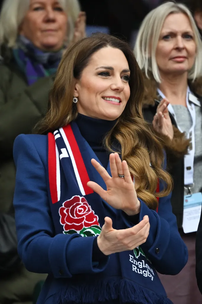 Catherine, Princess of Wales, smiles while wearing a navy blue scarf with an embroidered red rose and the words