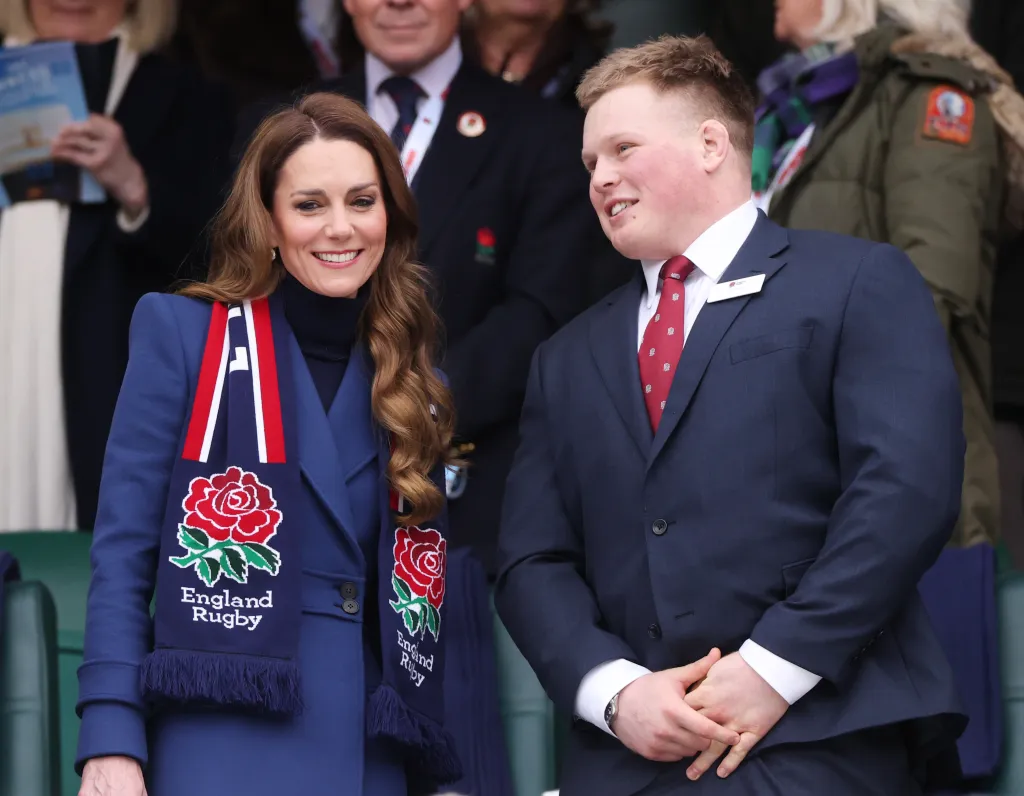 Catherine, Princess of Wales, wearing a blue coat and England Rugby scarf, smiles next to Fin Baxter at a rugby match.