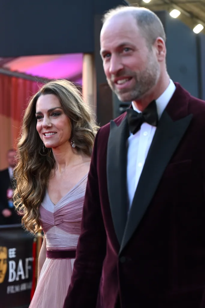 Catherine, Princess of Wales, in a pink gown, and Prince William, Prince of Wales, in a maroon velvet suit, walk together at the EE BAFTA Film Awards.