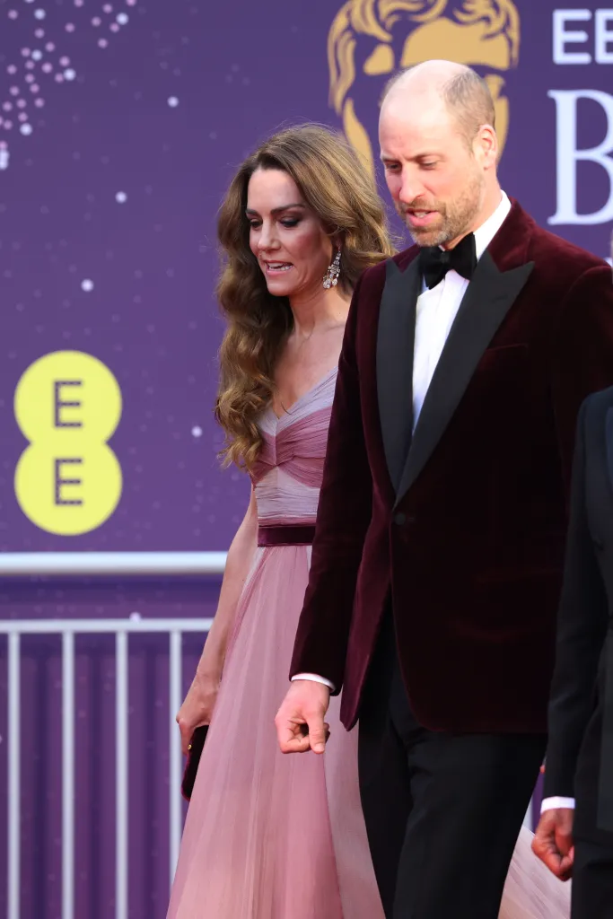 Catherine, Princess of Wales, in a pink gown, and Prince William, in a burgundy velvet tuxedo, walking on a purple carpet at the BAFTA Film Awards.