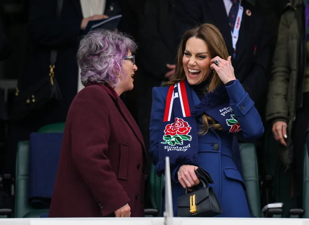 Catherine, Princess of Wales, in a blue coat and England Rugby scarf, laughs while talking to a woman with purple hair.