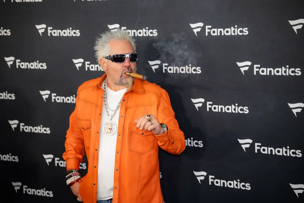 Guy Fieri, wearing an orange jacket and sunglasses, smokes a cigar at the Fanatics Super Bowl Party.
