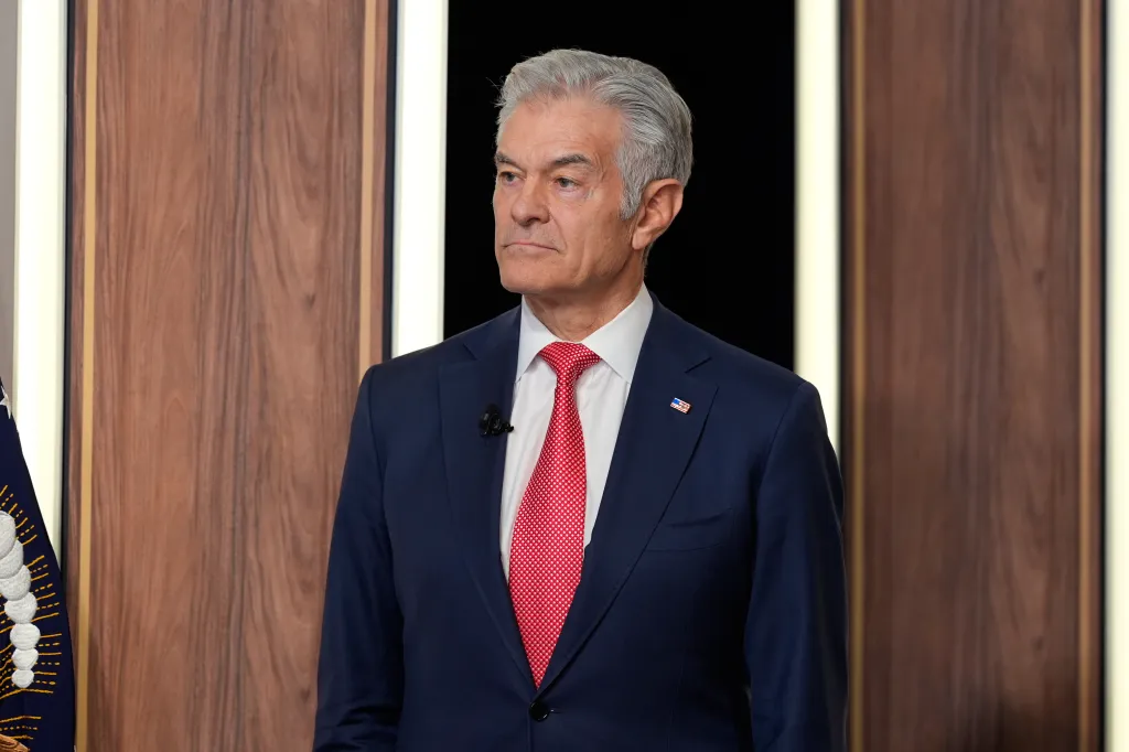 Dr. Mehmet Oz, wearing a suit and red tie, stands listening.