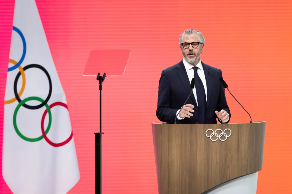 Casey Wasserman speaking at a podium with the Olympic rings logo, against a bright red background.