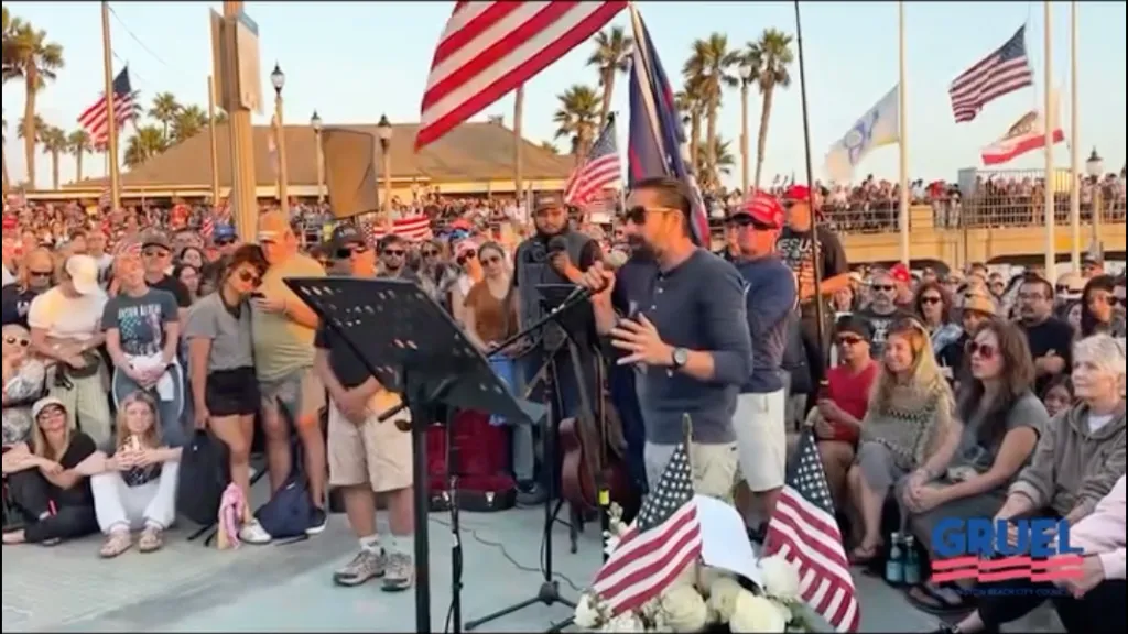 Chef Andrew Gruel speaking into a microphone in front of a crowd with American flags.