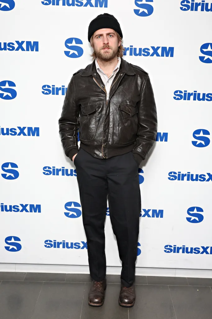 Christian Lee Hutson in a black beanie and brown leather jacket, standing in front of a white wall with blue SiriusXM logos.