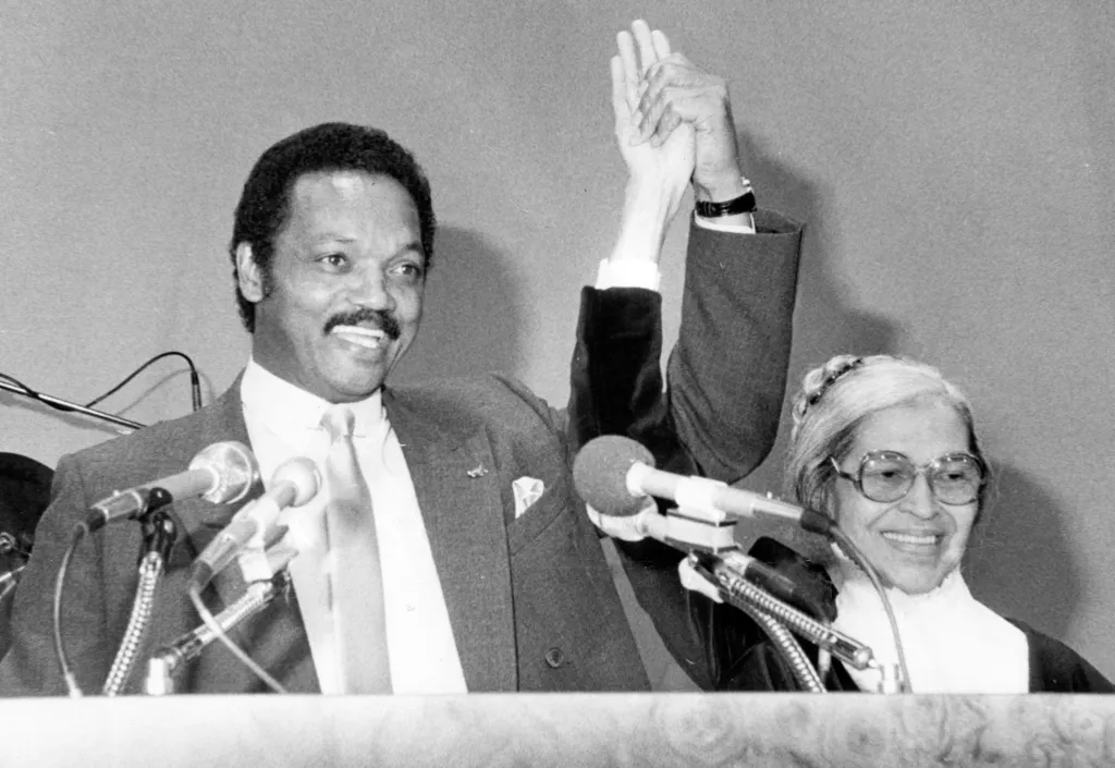 Black and white photo of Jesse Jackson and Rosa Parks raising joined hands triumphantly.