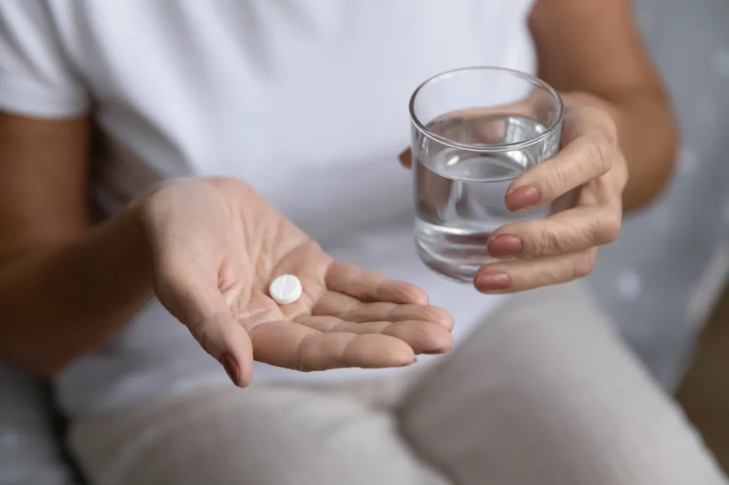 A person holding a white pill in one hand and a glass of water in the other.