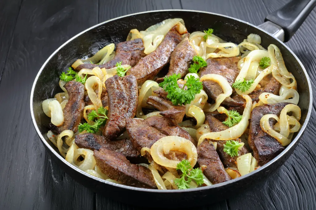 Fried calves liver pieces with onion rings in a skillet.