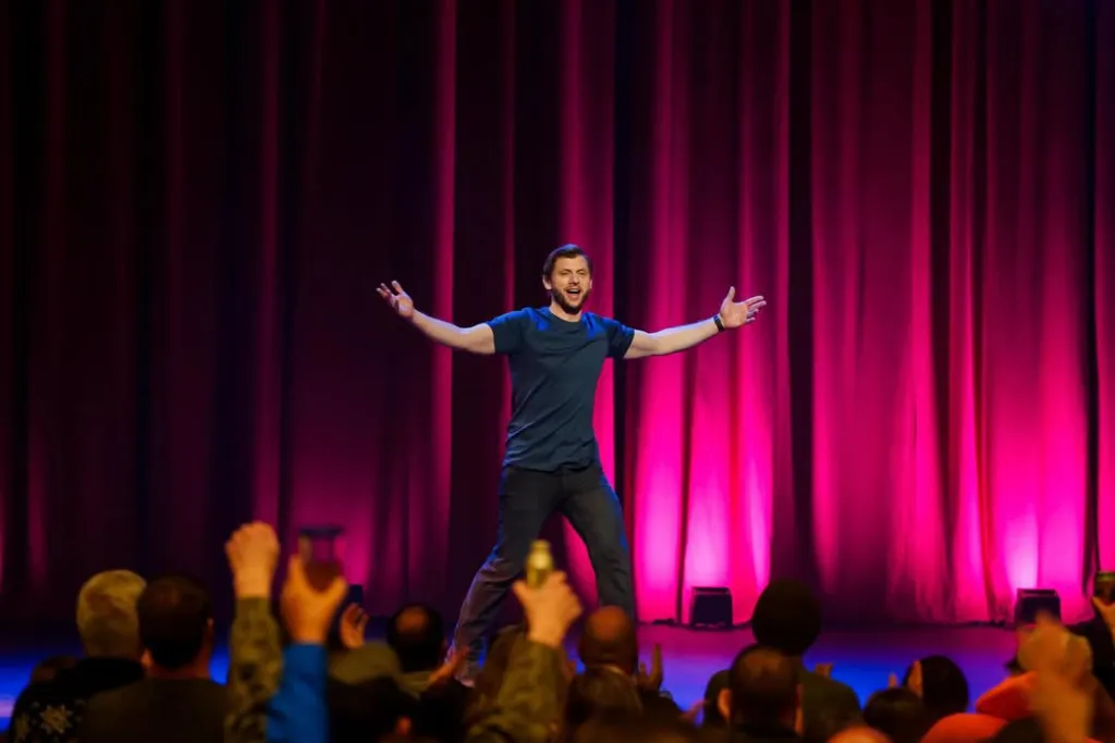 Comedian Charlie Behrens performs on stage with his arms outstretched.