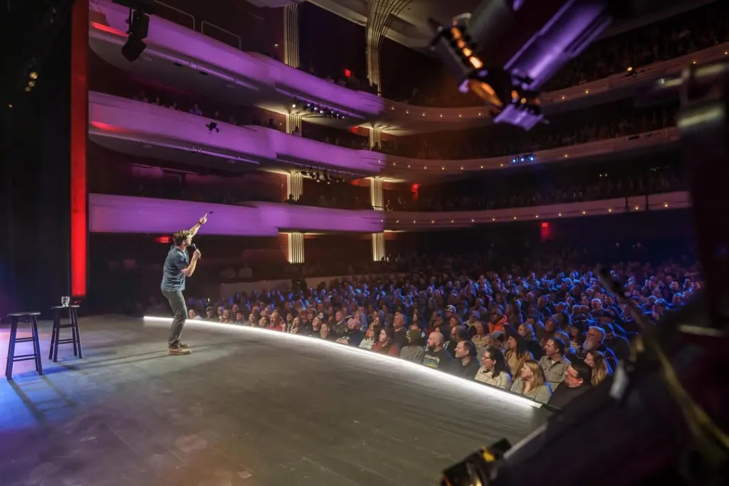 Comedian Charlie Behrens performs on stage, gesturing to the audience.