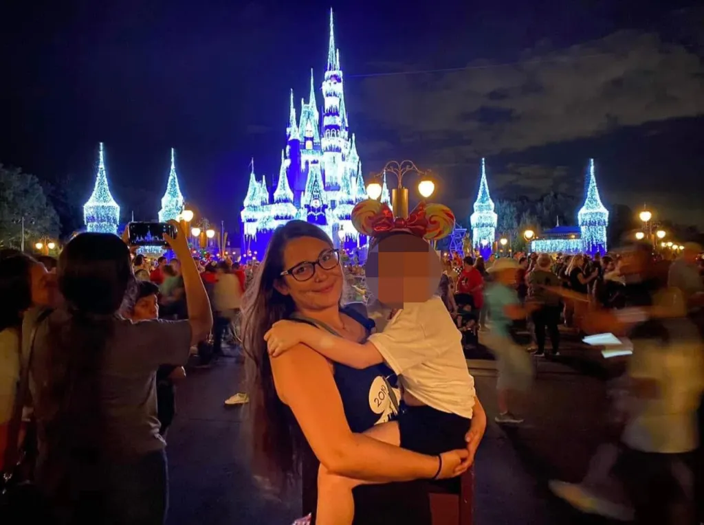 Courtney Drysdale holding a child with a blurred face in front of a brightly lit Cinderella Castle at night.