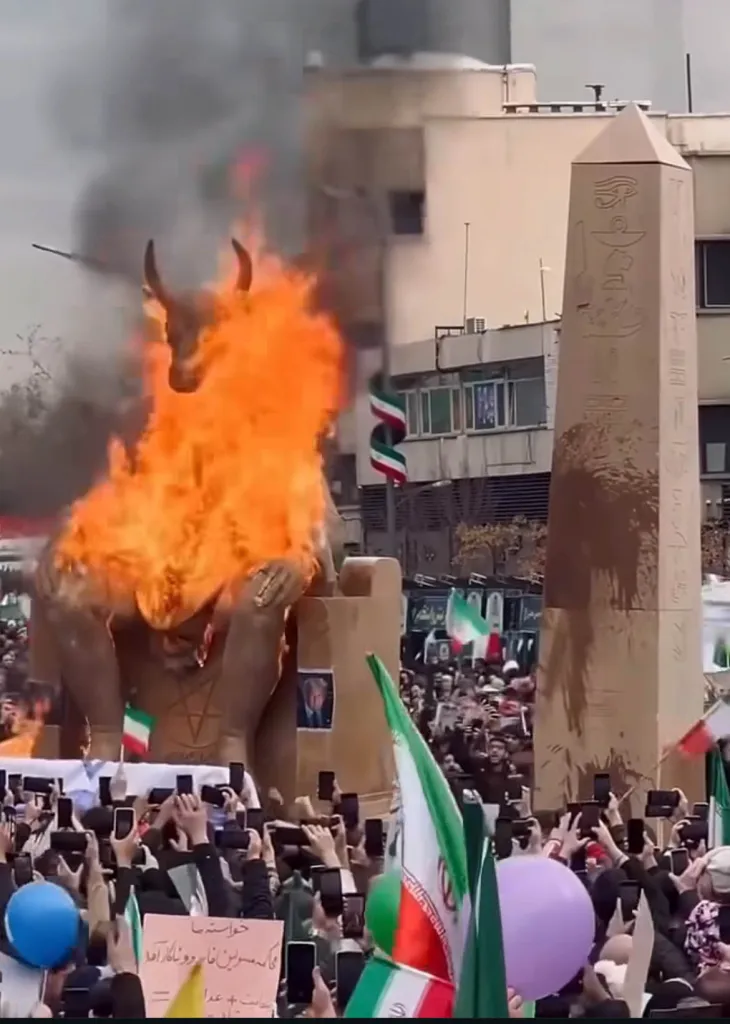 A large golden statue of a bull, representing Ba'al, burns fiercely in a crowd of protestors holding up phones and Iranian flags.