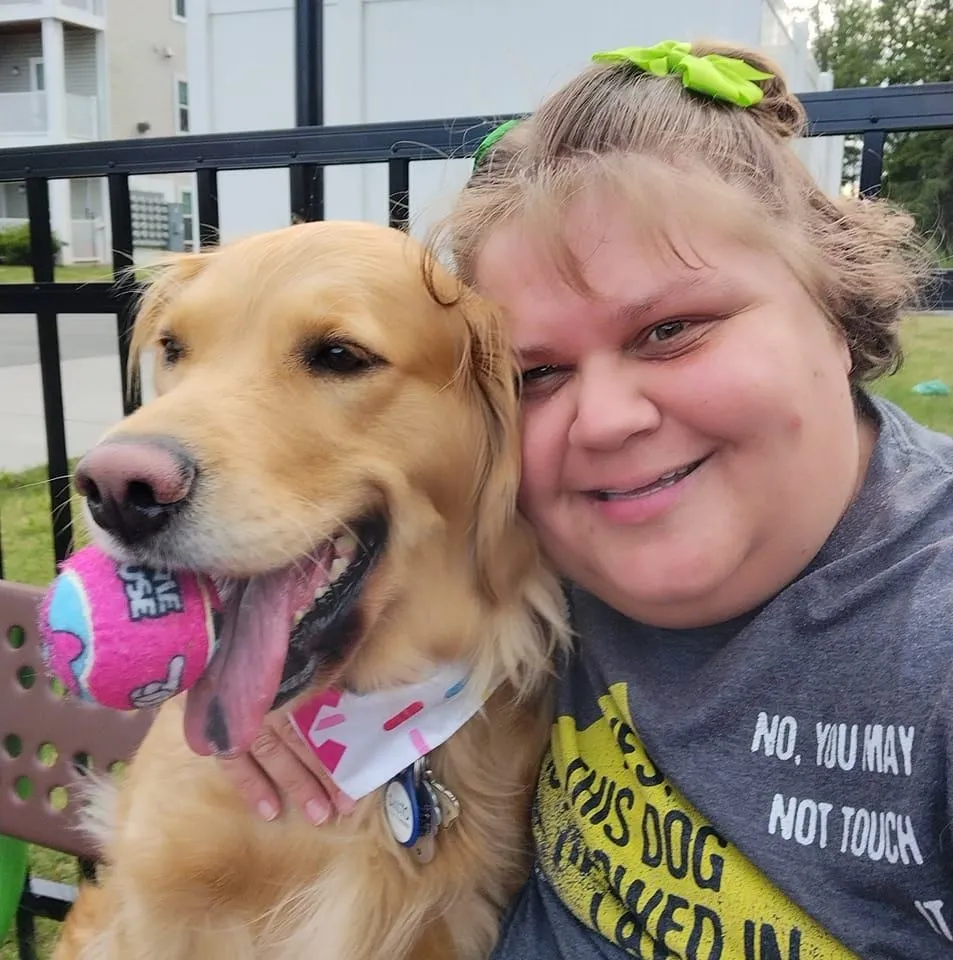 Crystal Pratt smiles with her service dog, a golden retriever, who holds a pink tennis ball in its mouth.