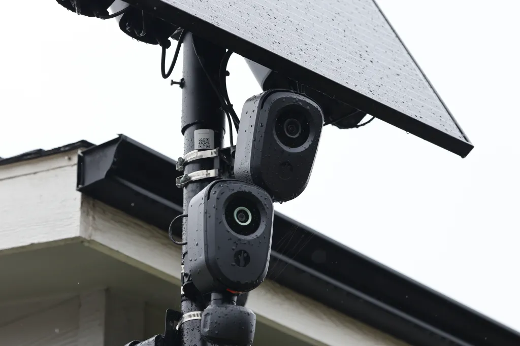 Two outdoor security cameras and a small solar panel are attached to a pole, with a house visible in the background.