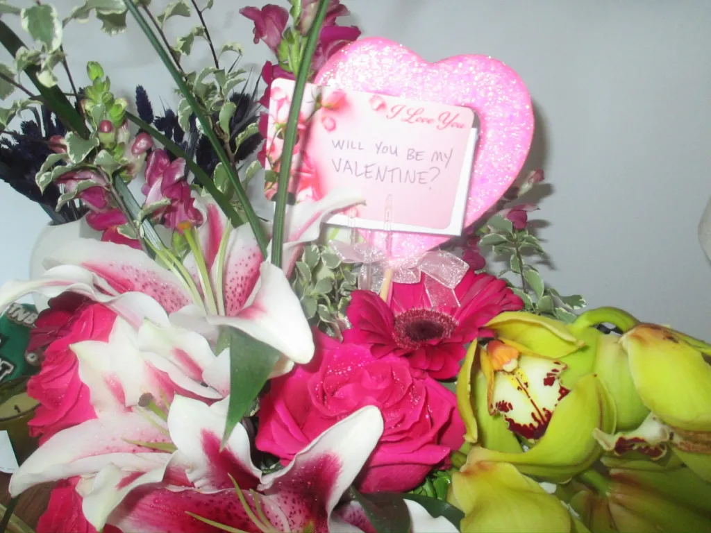 A bouquet of various flowers including lilies, roses, and orchids, with a glittery pink heart-shaped card that reads,
