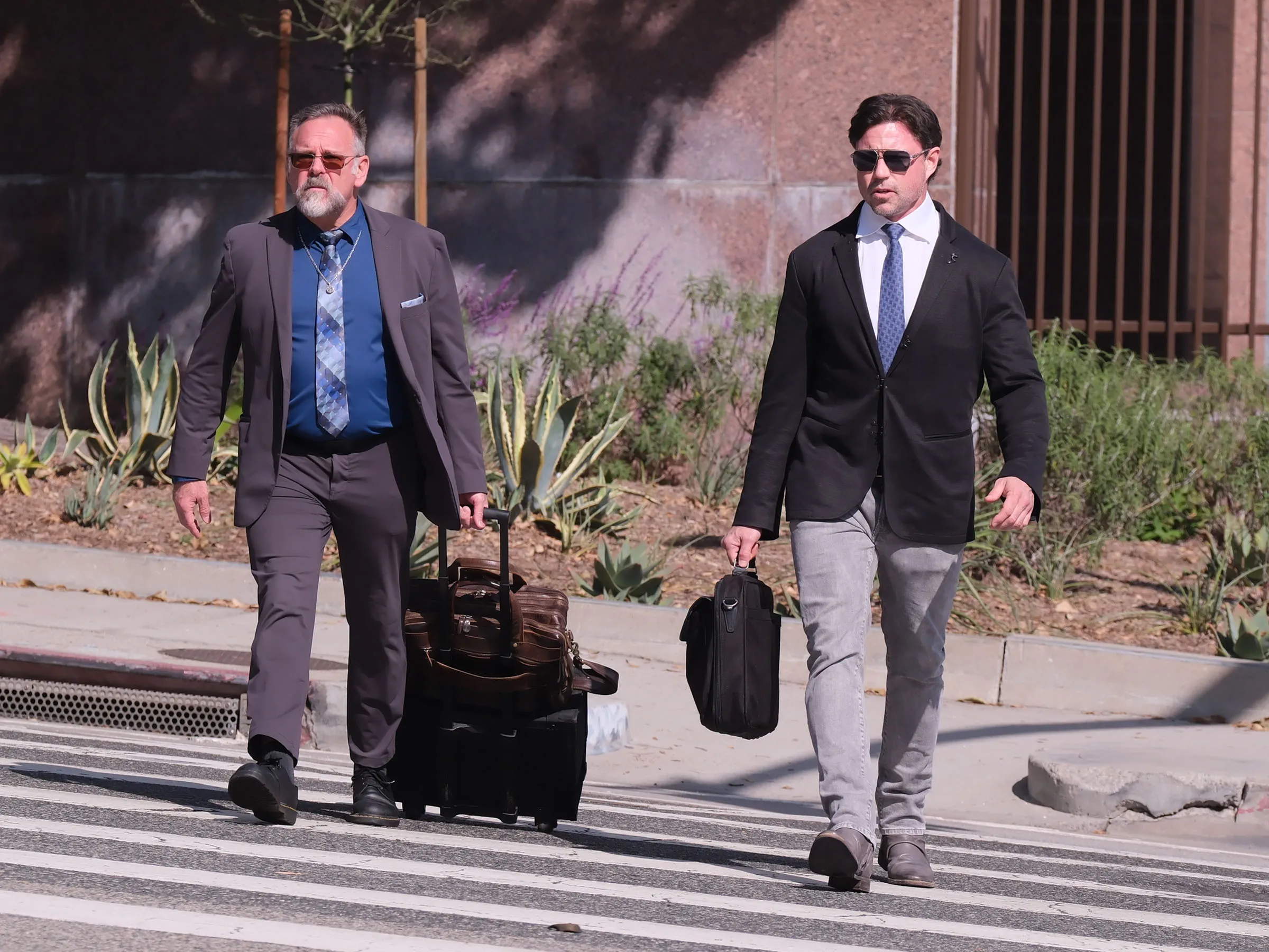 Aaron Phypers in a black suit, white shirt, and blue tie, walking with a briefcase, beside another man wearing a gray suit, blue shirt, and blue tie, pulling a rolling suitcase.