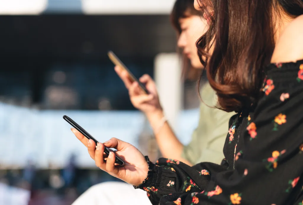 Two young women sitting side by side and looking at their smartphones.