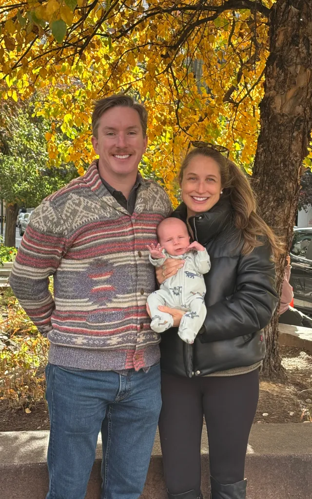 A man, woman, and baby stand smiling in front of a tree with yellow leaves.