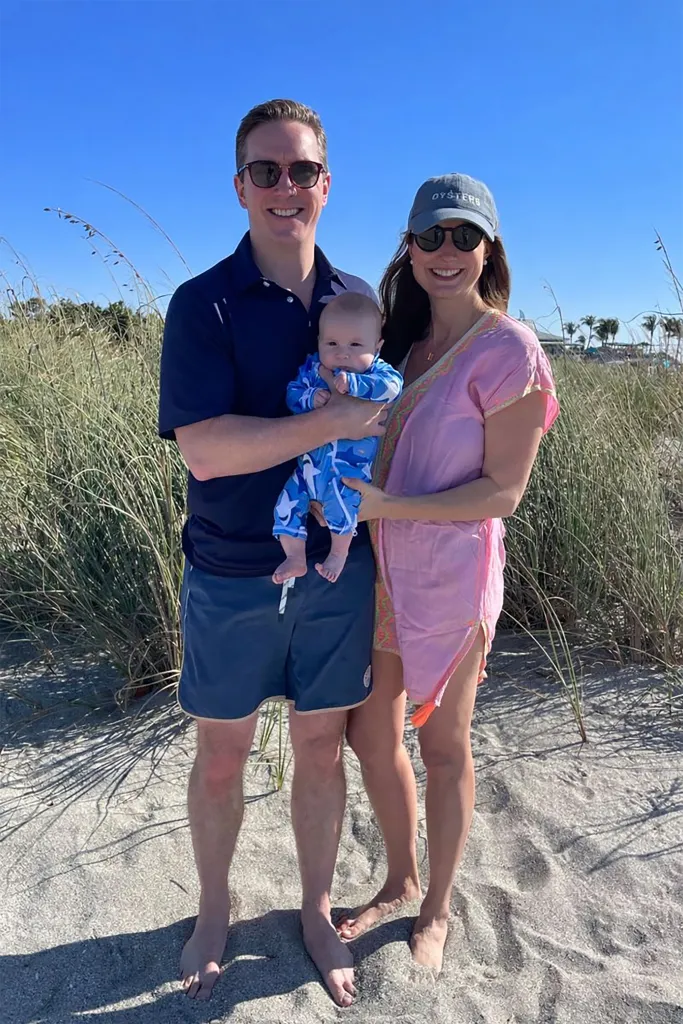 A man, woman, and baby stand on a sandy beach in front of tall grass.