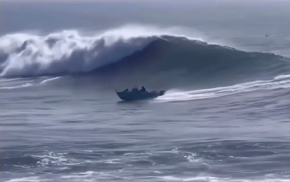 A small boat with passengers rides a large ocean wave.
