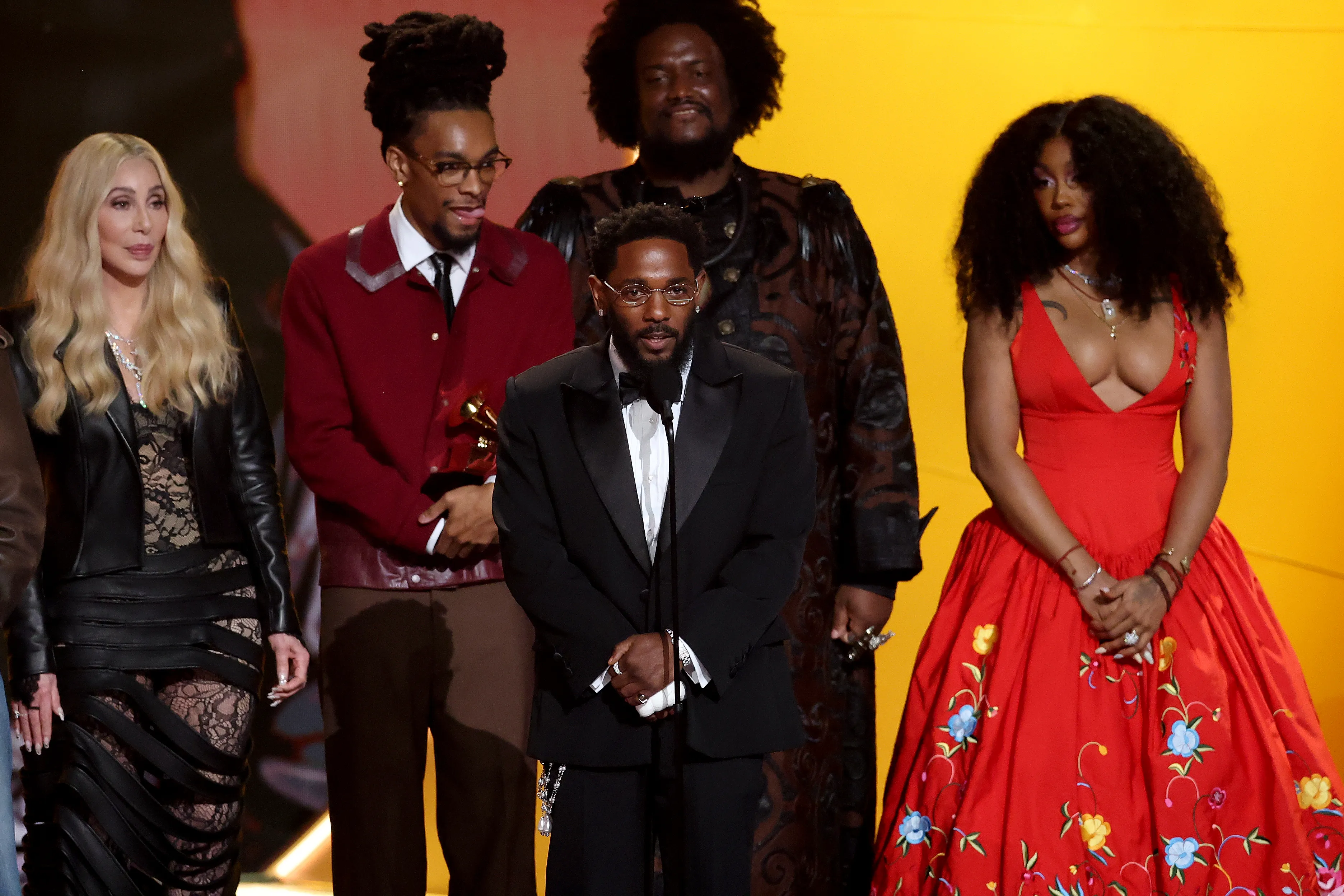 Kendrick Lamar, wearing a black tuxedo, accepting the Record of the Year award alongside SZA, wearing a red gown, and other collaborators.
