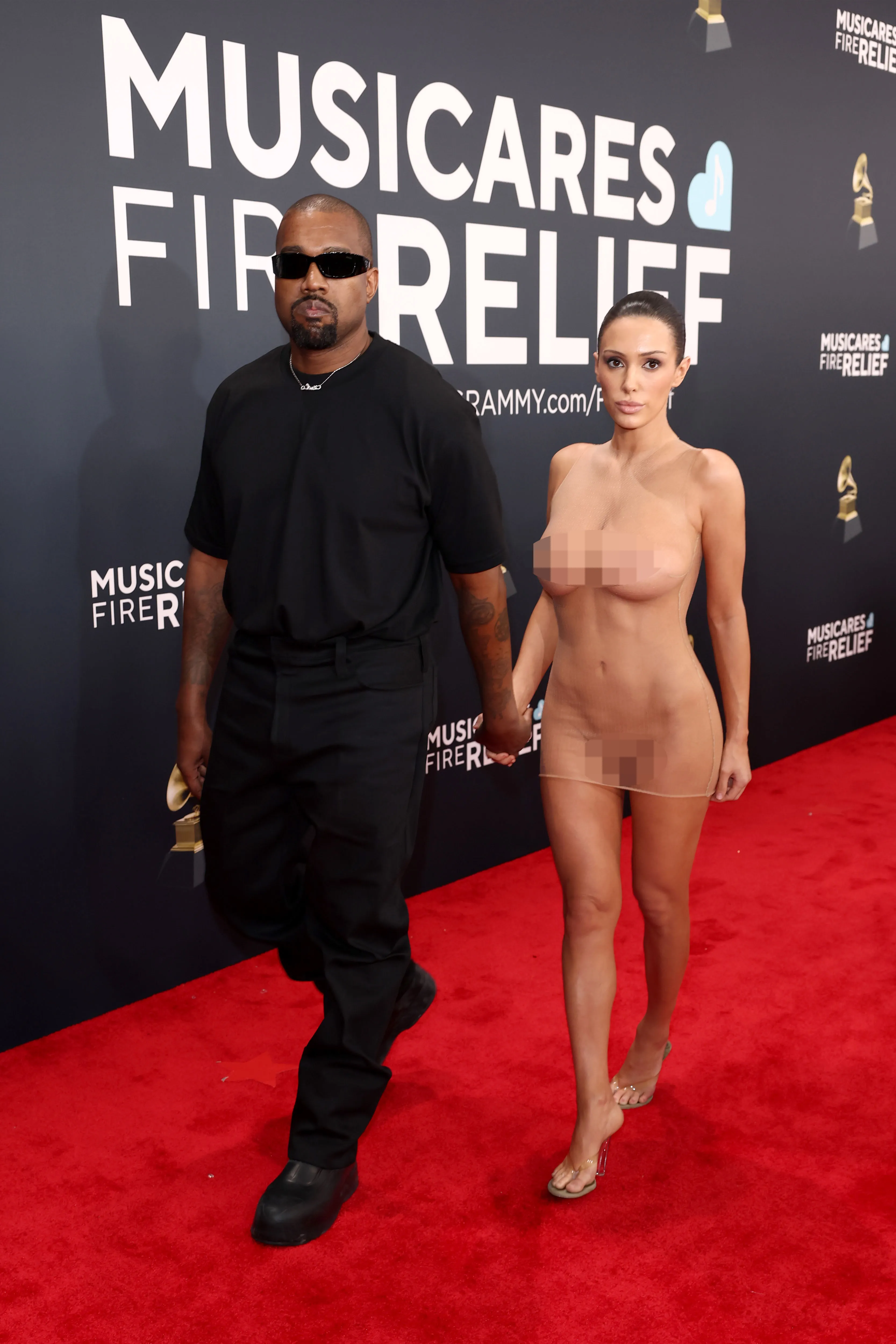 Kanye West, in a black shirt and pants, walks hand-in-hand with Bianca Censori, who is wearing a sheer, skin-toned dress, down a red carpet.