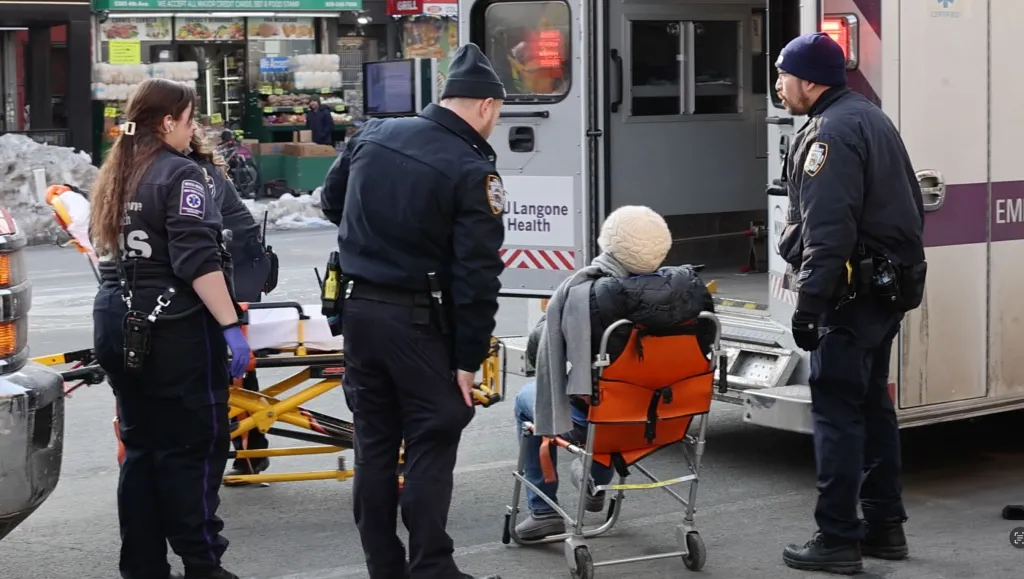 An EMT and two NYPD Transit officers assist a person in a transport chair next to an ambulance.