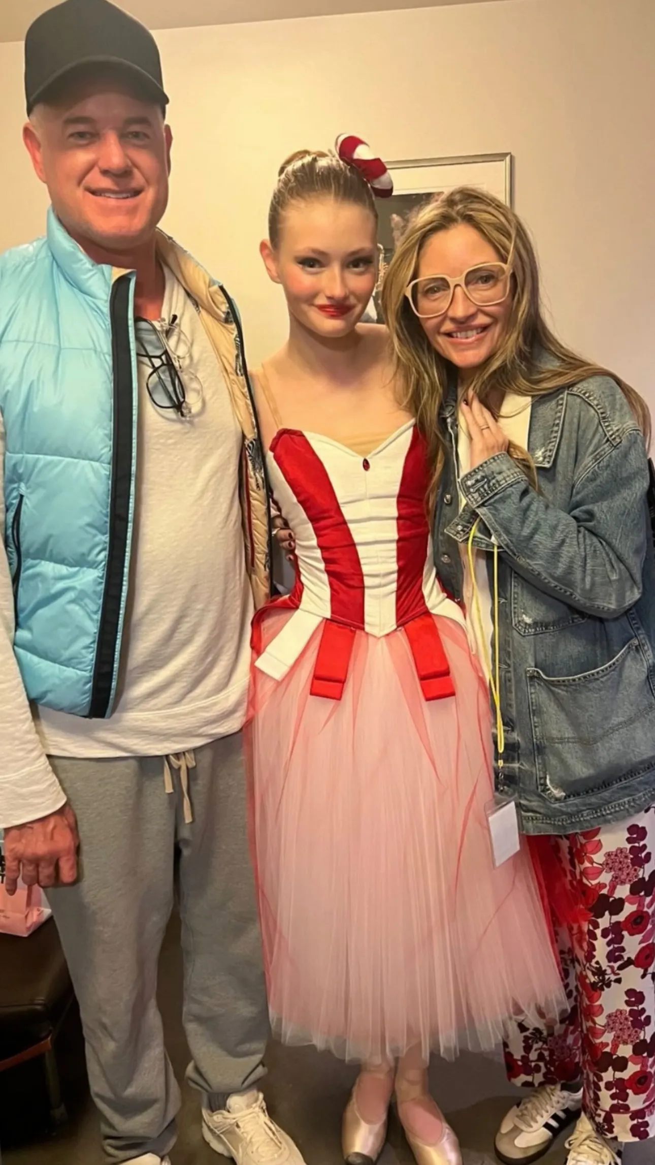 A man in a black cap and blue vest, a girl in a red and white ballet costume, and a woman in glasses and a denim jacket stand together, smiling.