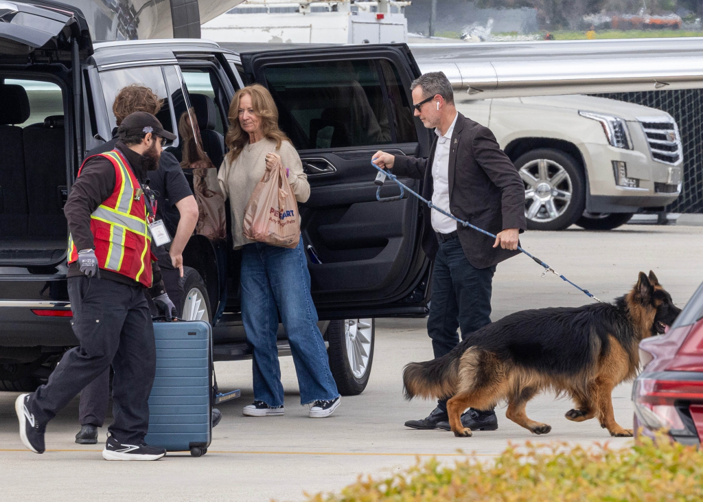 Lisa Mudd Sweeney exiting an SUV with a dog being led on a leash by a man, and a luggage handler pulling a suitcase.