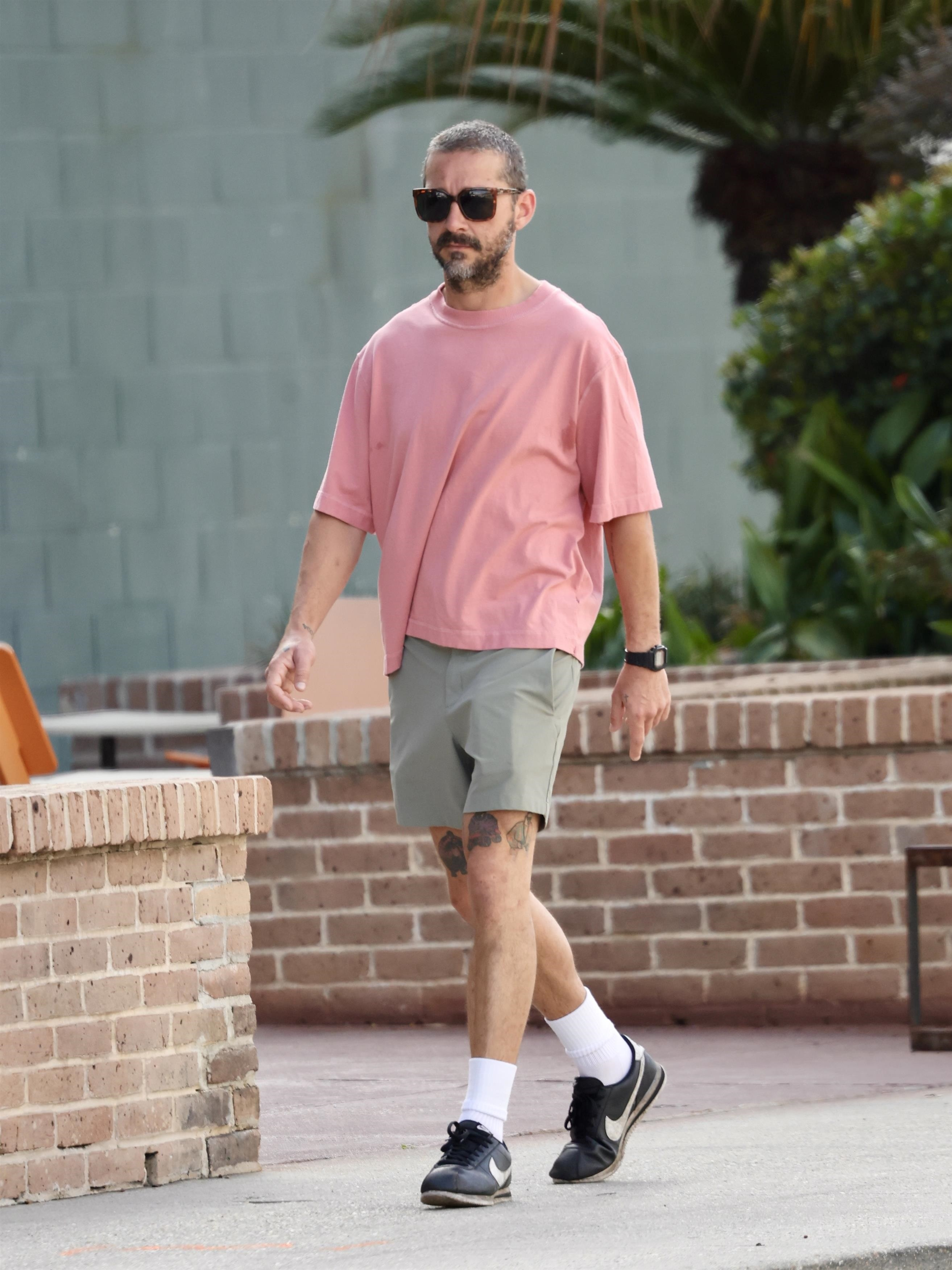 Shia LaBeouf walking outdoors in a pink shirt, gray shorts, and sunglasses.