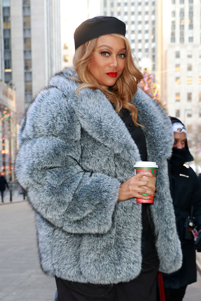 Tyra Banks in a gray fur coat and black beret, holding a Starbucks coffee cup.