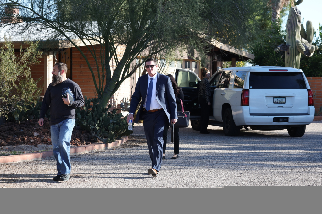 Law enforcement officials, including men in a suit and casual clothes, walking away from a white SUV parked in a gravel driveway.