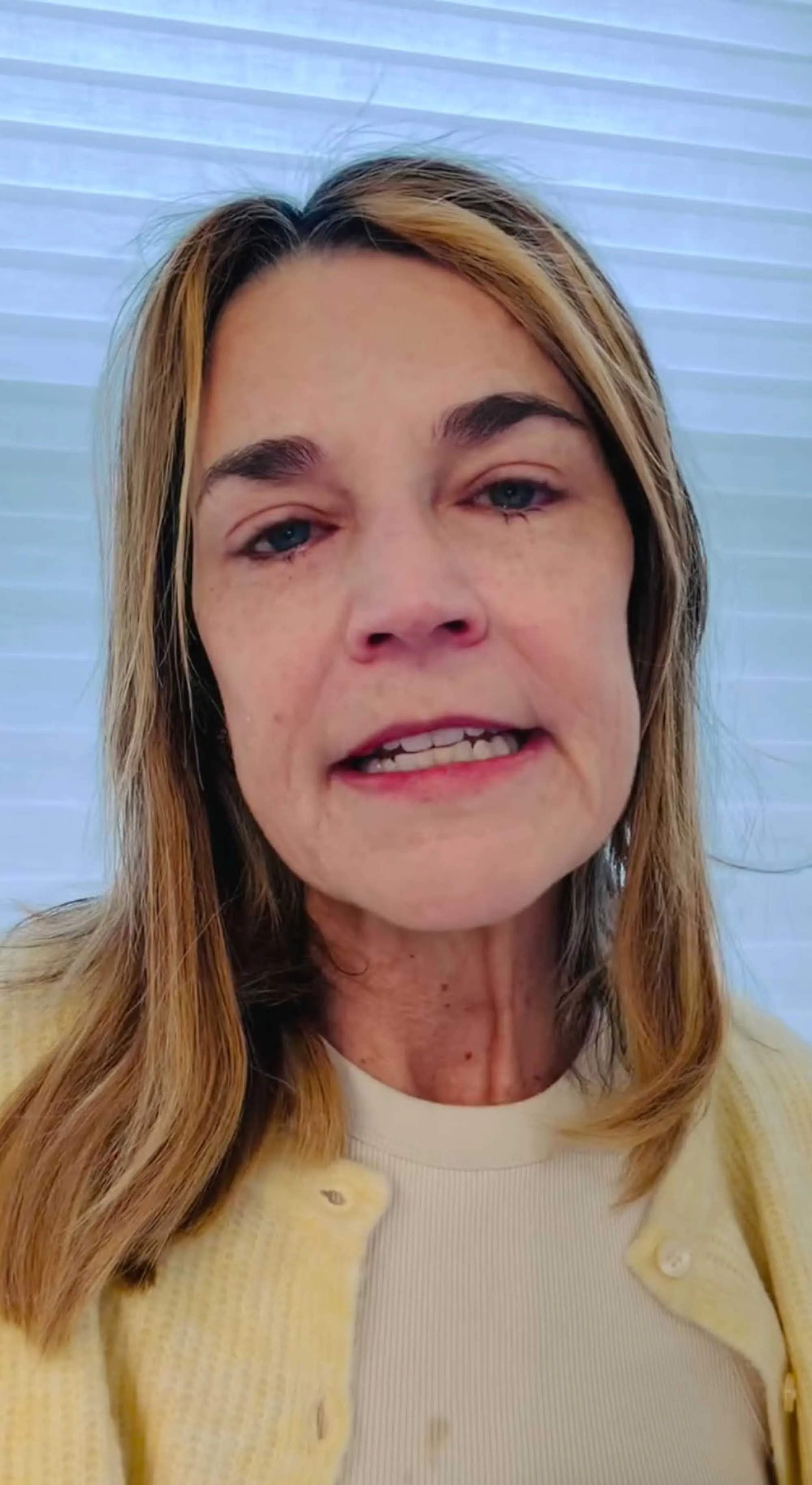 A light-skinned woman with blonde hair, wearing a white shirt and a yellow cardigan, speaks directly to the camera.