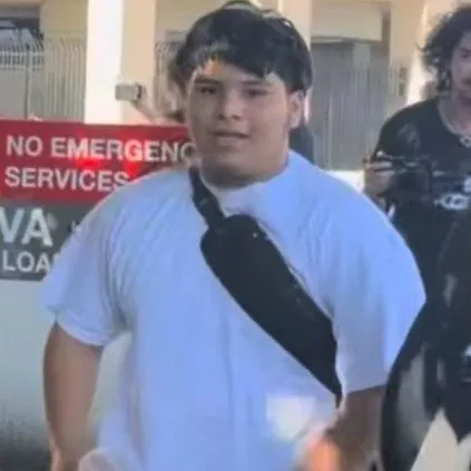 A person in a white T-shirt and khaki pants walks with a drink in hand.