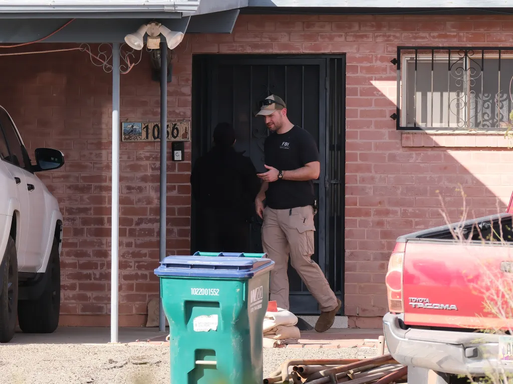 An FBI agent wearing a black t-shirt and tan pants speaks to someone at the door of a brick house.