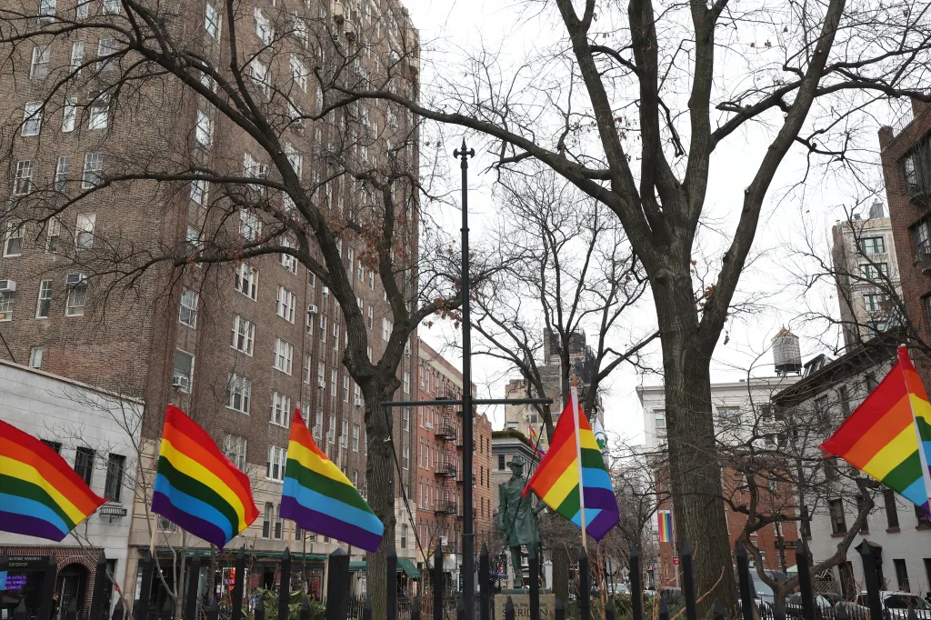 Rainbow flags fly at the Stonewall National Monument, with tall buildings and bare trees in the background.