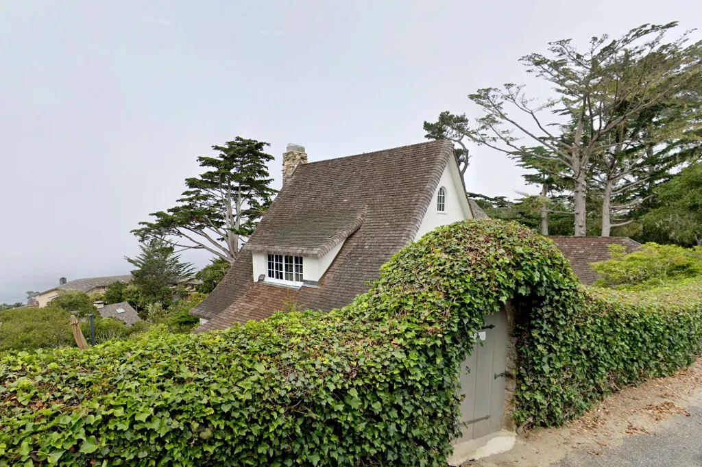 Flor Mora's house at 110 Pine Way, Carmel, CA, featuring a brown shingled roof, white dormer windows, and a dense ivy hedge with an arched entryway.