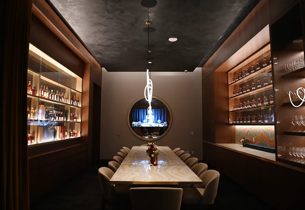 Dining room at Carversteak with a long marble table, displays of liquor bottles, and a large circular mirror reflecting a screen.