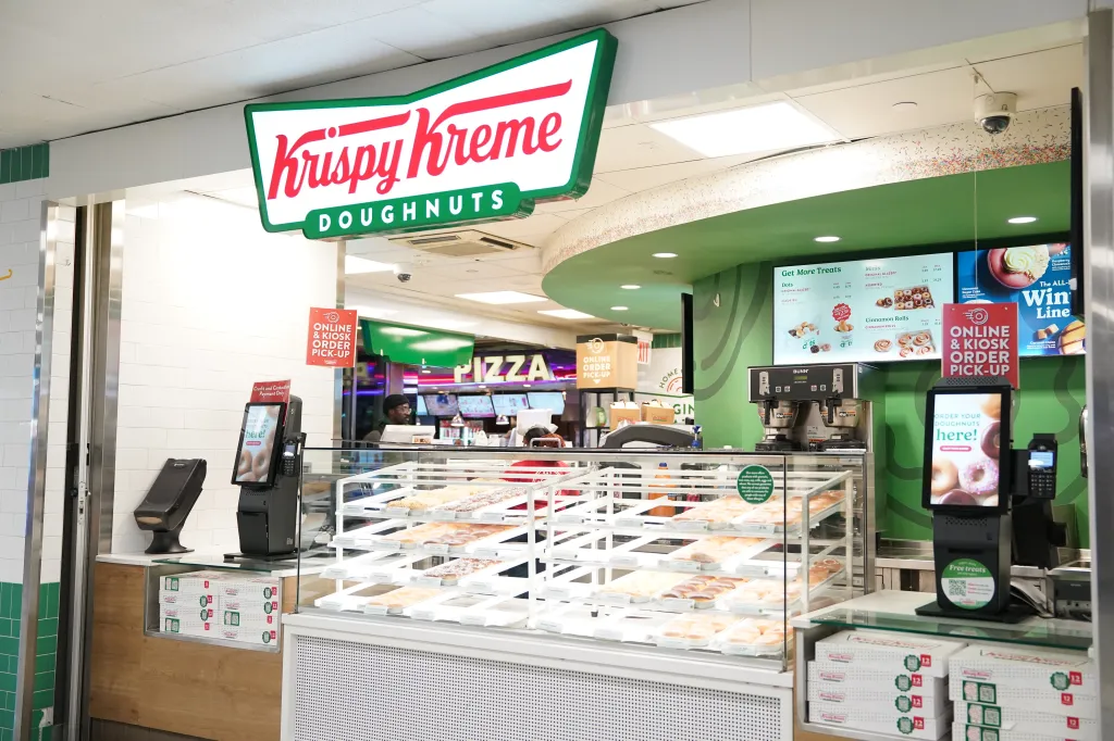 A Krispy Kreme Doughnuts store with a large display case of donuts, a digital ordering kiosk, and a large sign displaying the company's logo.