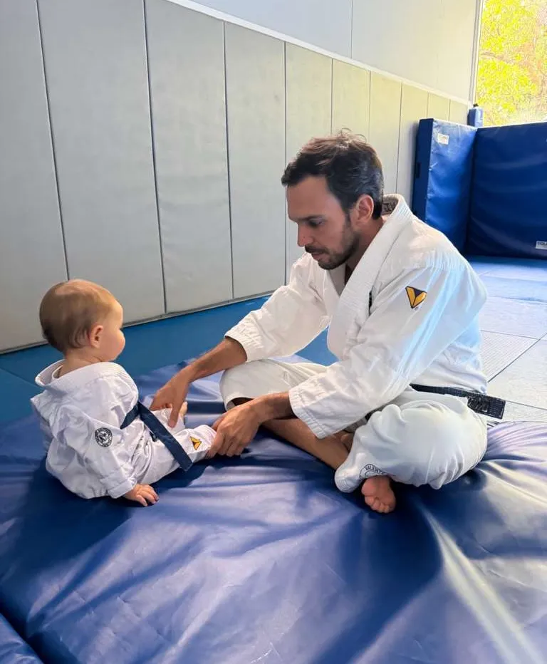 A man in a gi with a black belt sitting on a blue mat and adjusting a baby's gi.