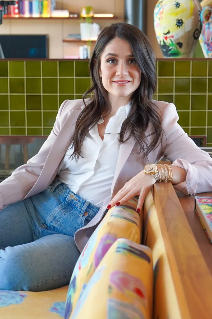 Gizem Ozcelik smiles, wearing a white shirt, light pink blazer, and blue jeans, sitting on a colorful couch.