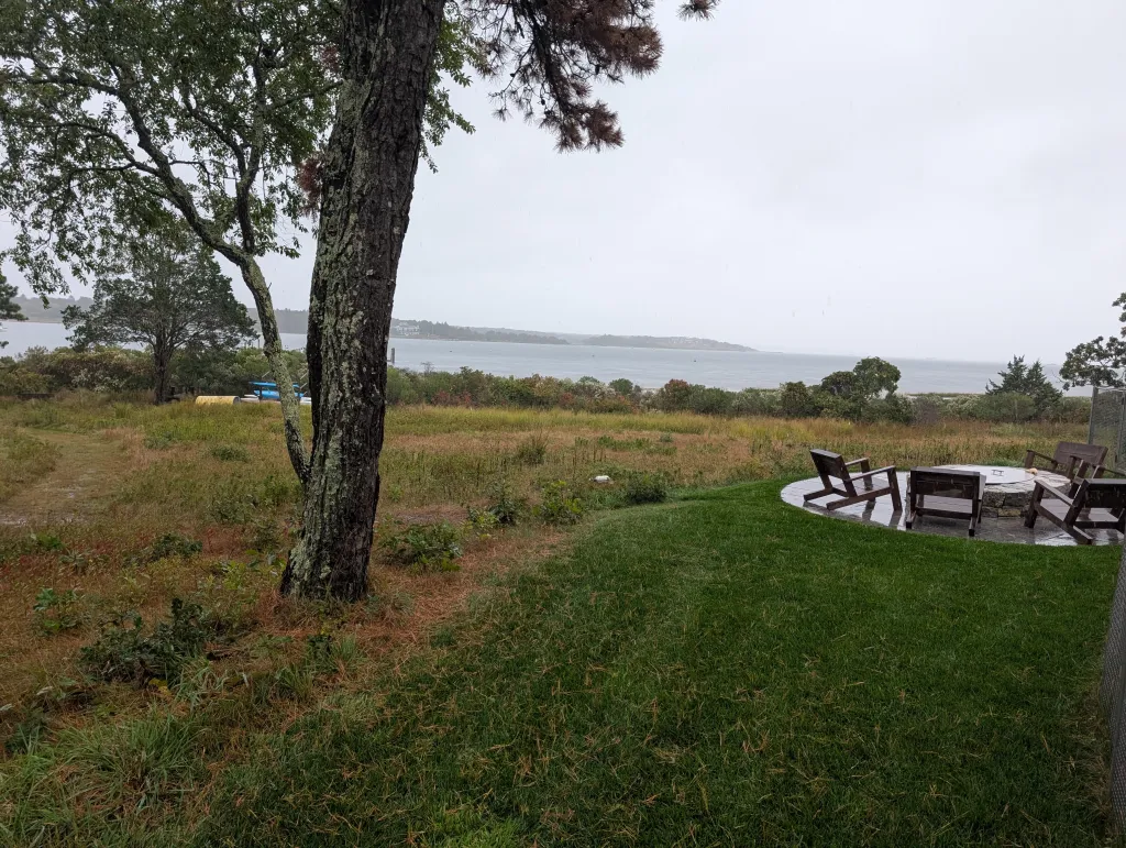 Outdoor patio with a stone fire pit and four wooden chairs facing a bay.