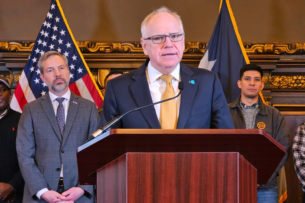 Gov. Tim Walz speaks at a news conference from behind a podium with several men standing behind him, including one to his left who has an American flag over his shoulder.