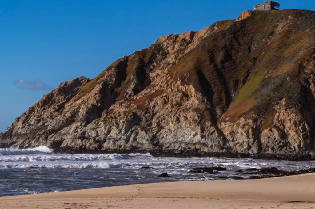 Gray Whale Cove State Beach in San Mateo County, California, showing waves crashing against the rocky shore below a high cliff with a building on top.