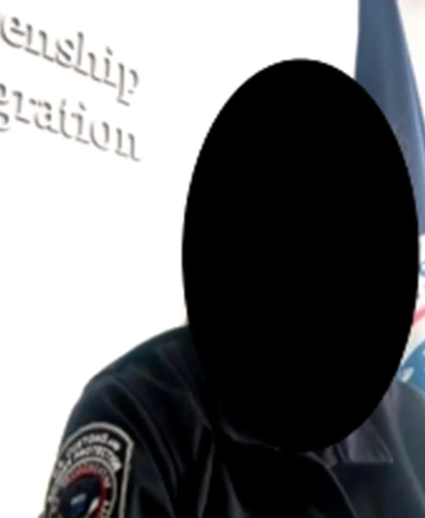 Cropped image of a person with their face obscured, wearing a uniform with a badge and standing in front of a sign that reads 