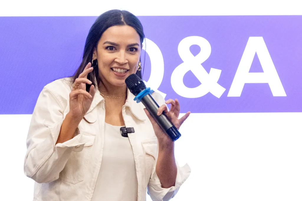 Rep. Alexandria Ocasio-Cortez speaking into a microphone with a Q&A sign behind her.