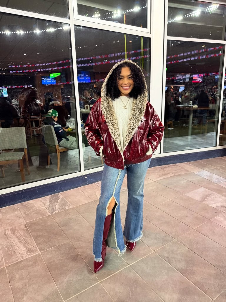 Victoria Gibson smiling in a red patent leather jacket with leopard print lining, ripped jeans, and red boots, standing in front of windows overlooking a stadium.