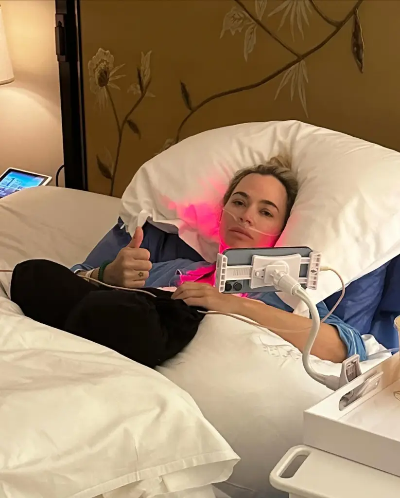Teddi Mellencamp recuperating in bed after surgery, giving a thumbs up.