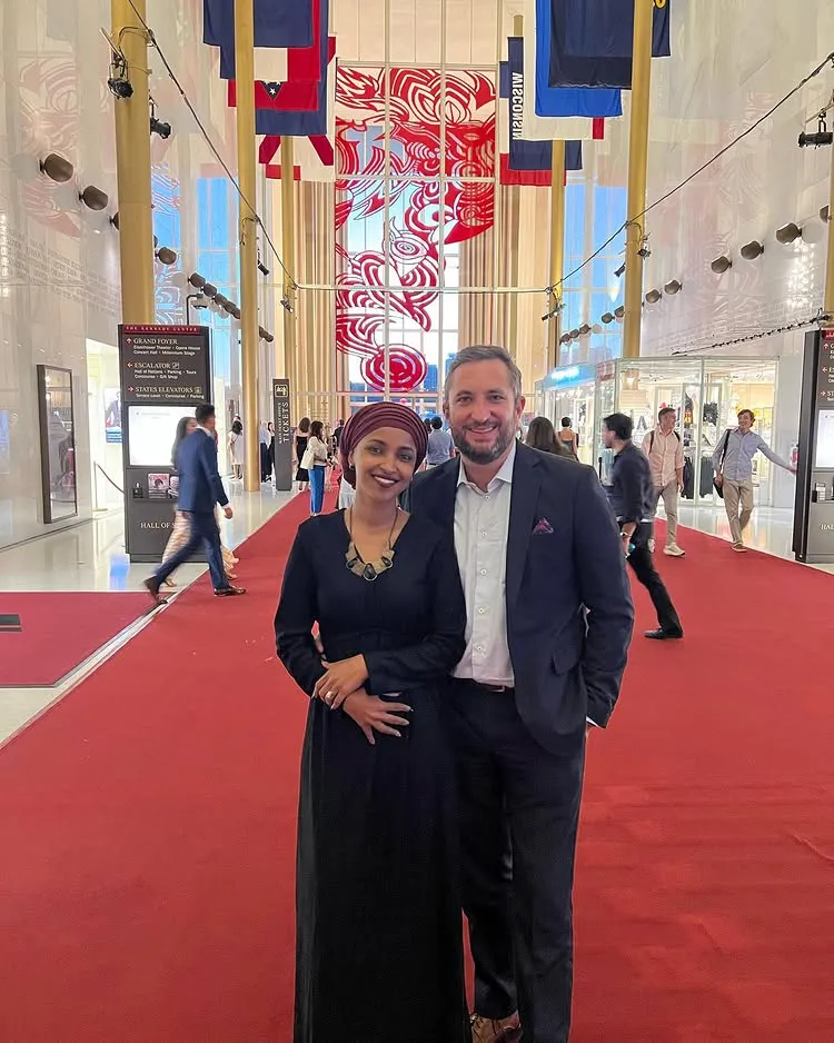Ilhan Omar and Tim Mynett posing on a red carpet in a grand hall.